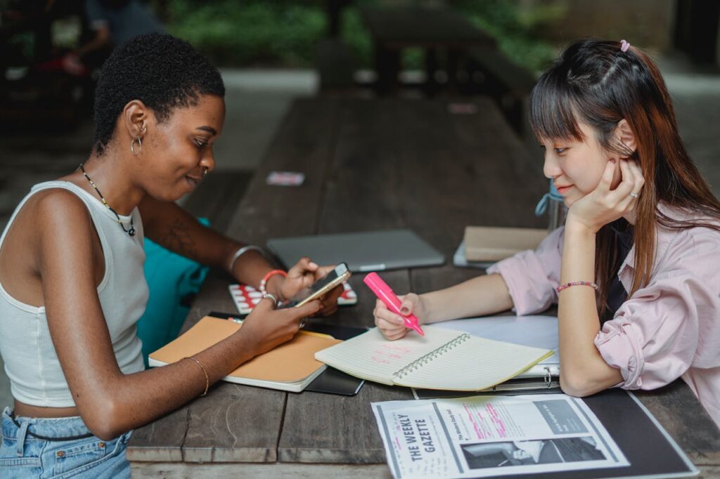 Two young women studying together outdoors, using a smartphone and notebook.